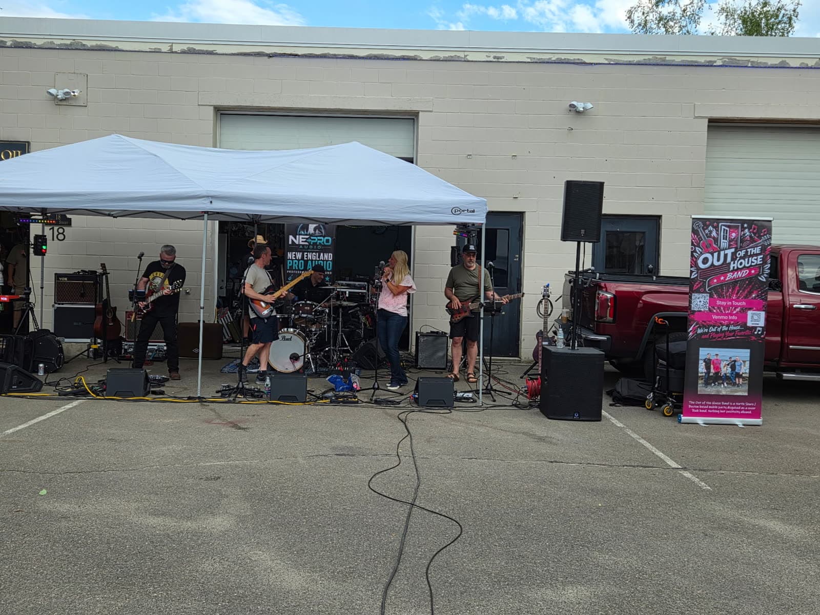 The band performing outdoors in daylight with the Out of the House Band banner